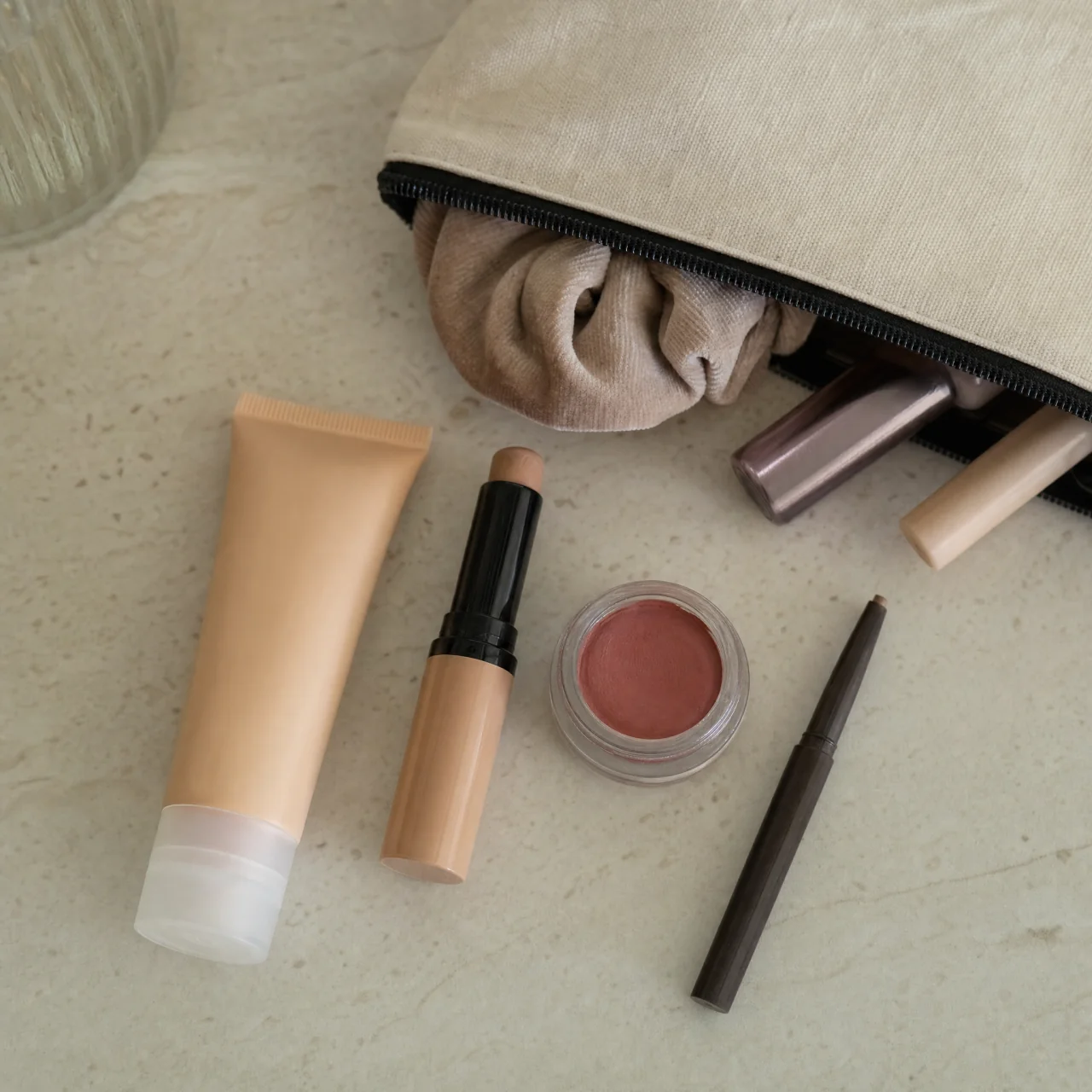 Flat-lay of a small curated makeup kit—tinted moisturizer, concealer stick, cream blush, brow pencil, mascara, and lip balm—arranged next to an open cloth pouch.