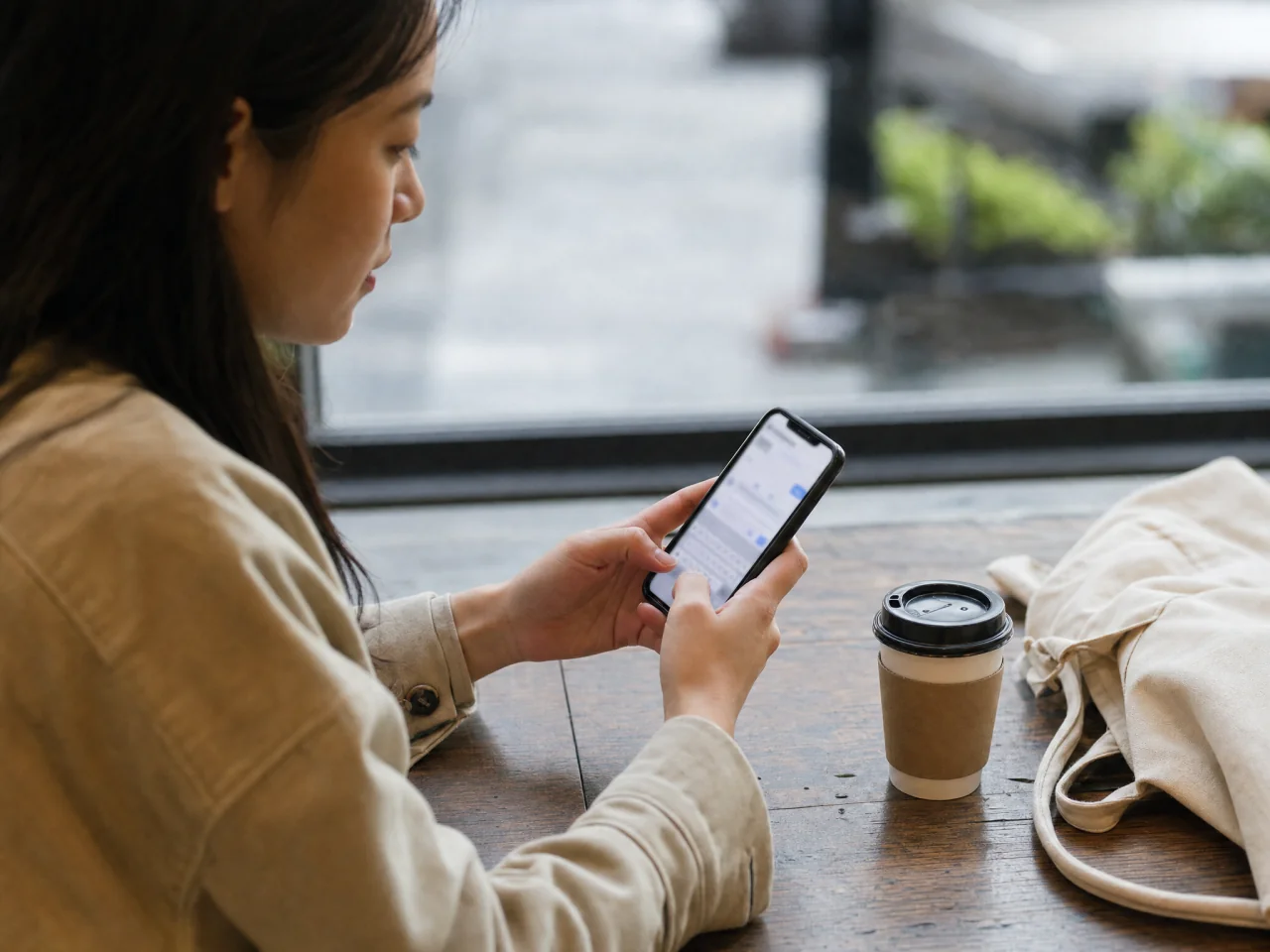 Commuter typing on a smartphone at a cafe table with a coffee cup and tote bag nearby