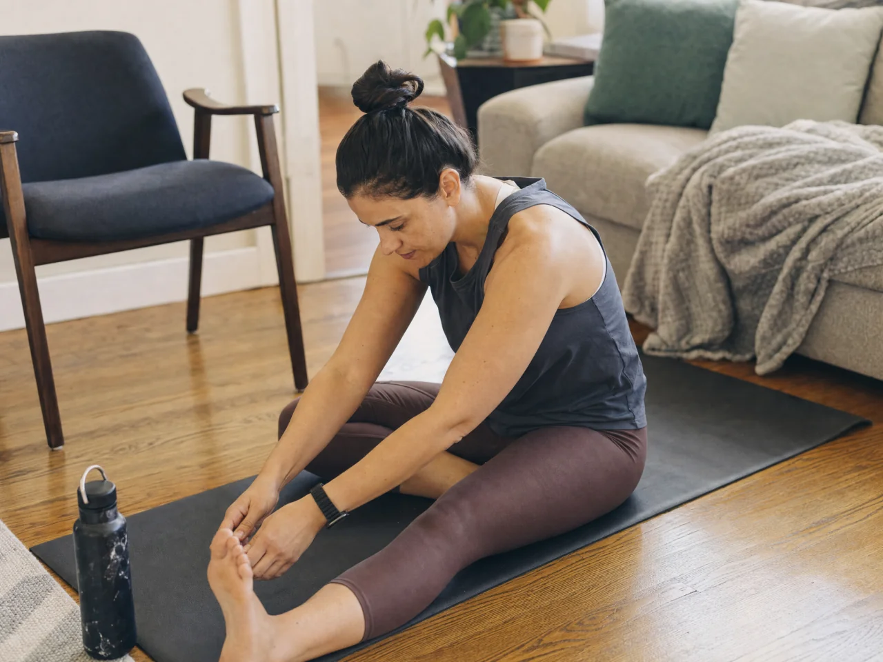 Person seated on an exercise mat doing a cool-down stretch with a chair and water bottle within reach.