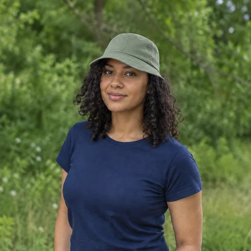 A woman with dark curls wears a sage-green bucket hat and navy shirt against softly blurred green foliage.