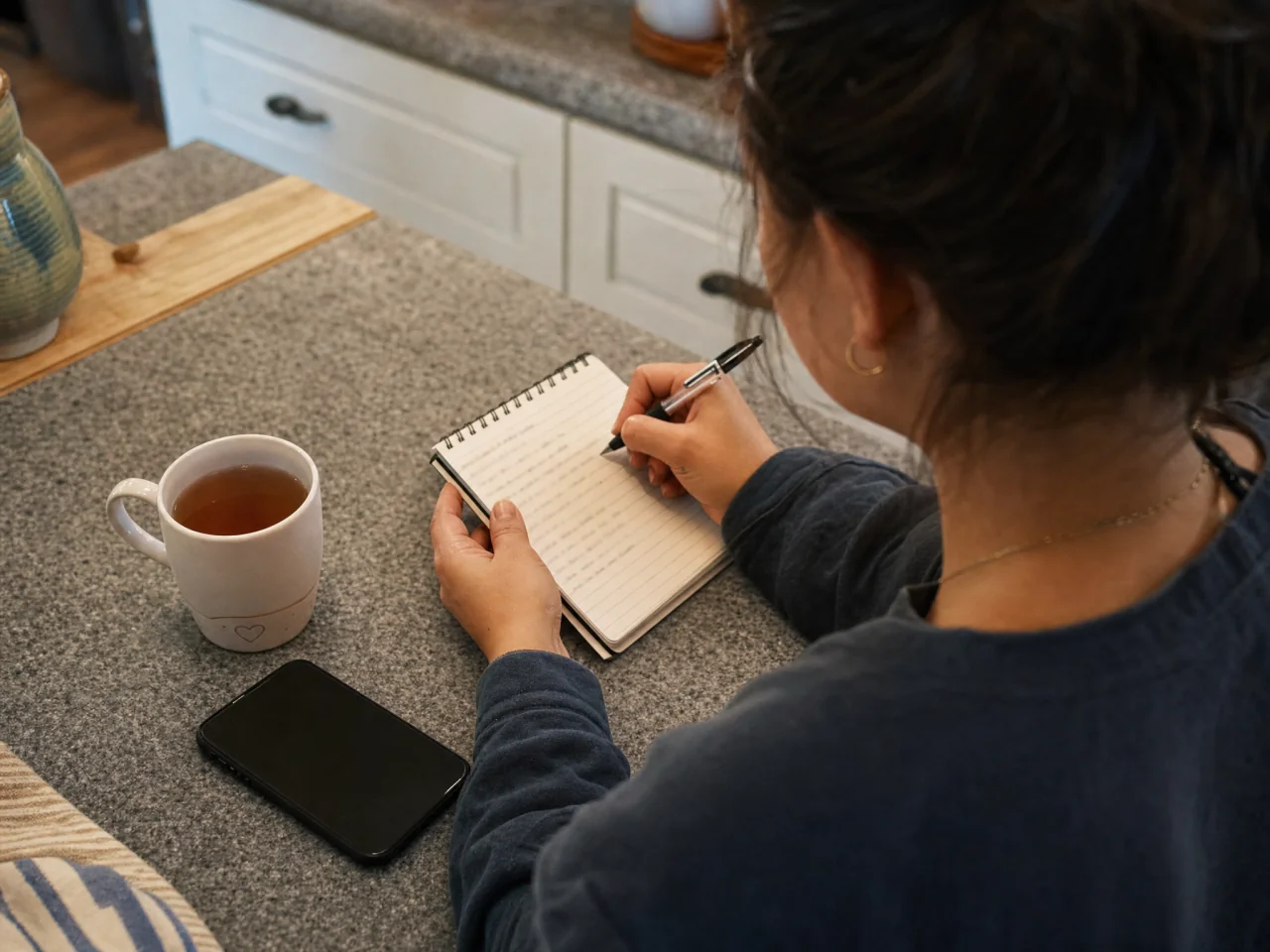 Person writing meal preferences in a pocket notepad at a kitchen counter; their hand partly covers the page so notes are not readable, with a mug of tea and a face-down phone nearby.