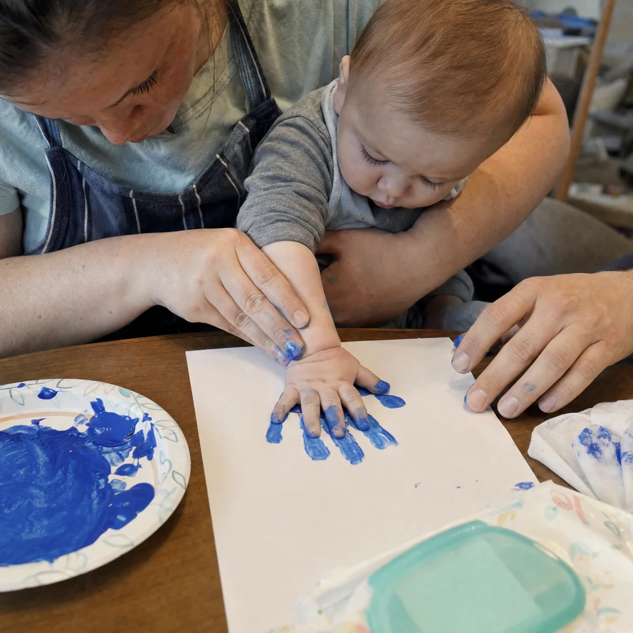 Caregiver guiding a baby's hand into washable paint and pressing it onto paper to make a handprint keepsake