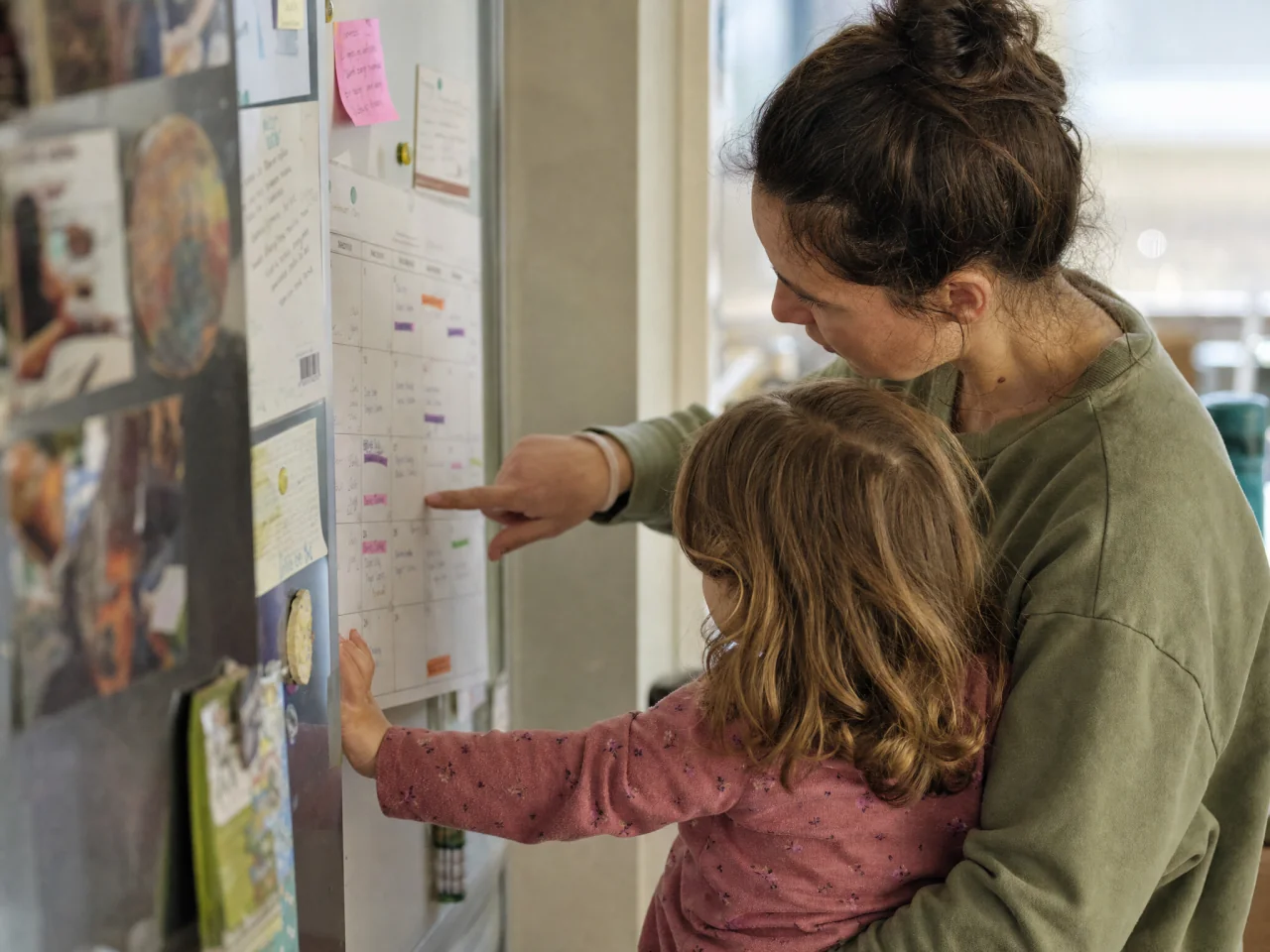 A parent checking a family calendar on the fridge while a small child stands nearby.