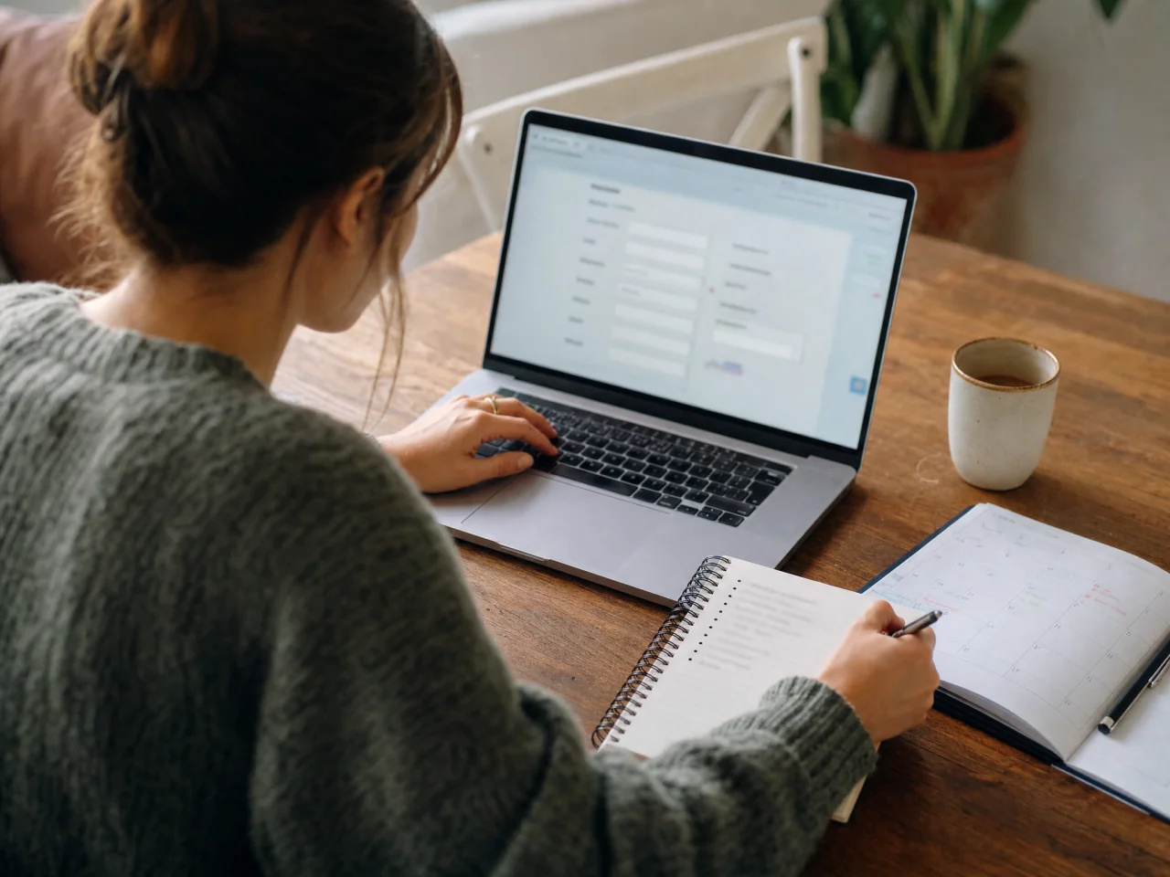 A person at a home table typing on a laptop and jotting notes in a notebook next to a calendar while planning a 60th birthday.