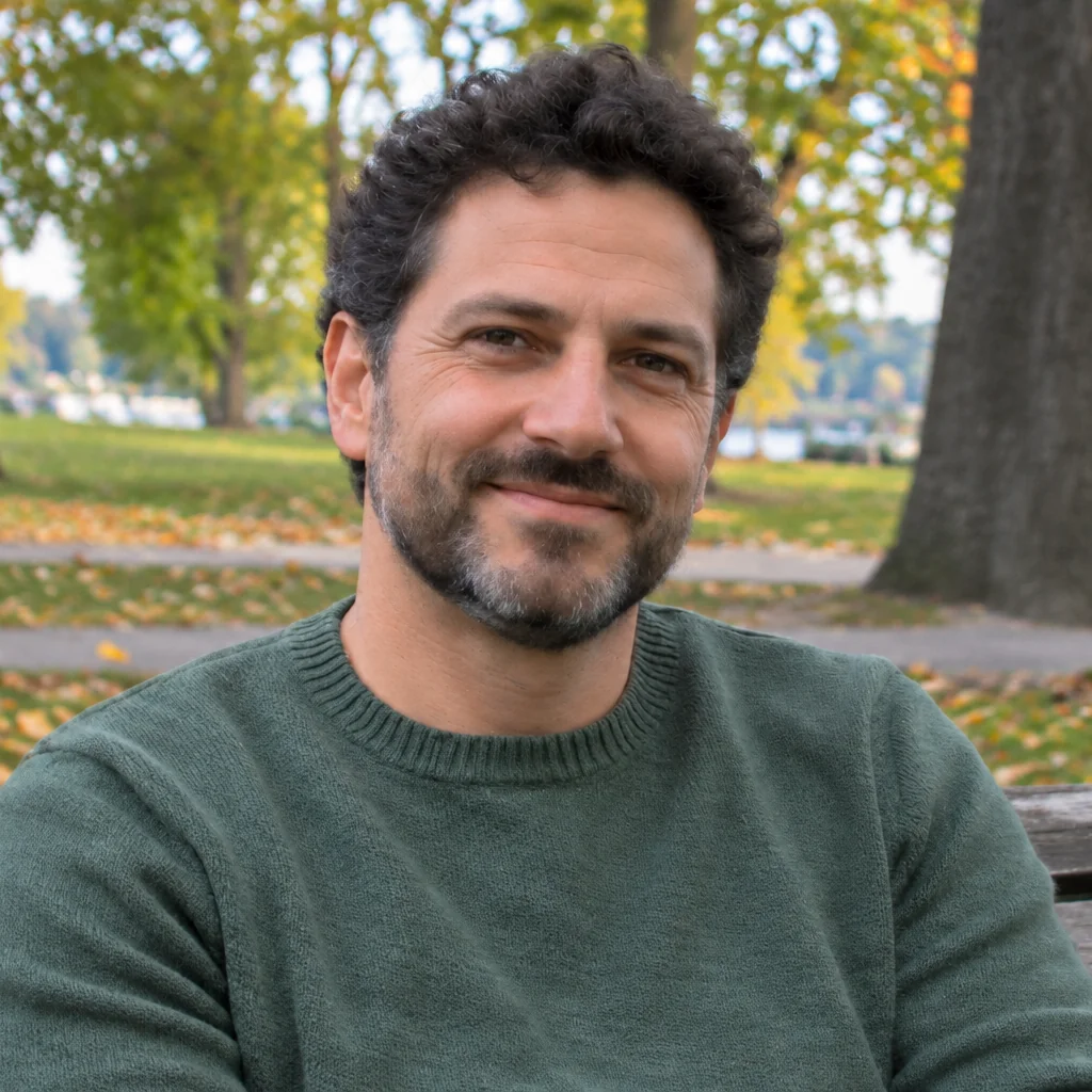 A smiling bearded man in a green sweater sits on a park bench, with clear facial detail and autumn trees behind him.