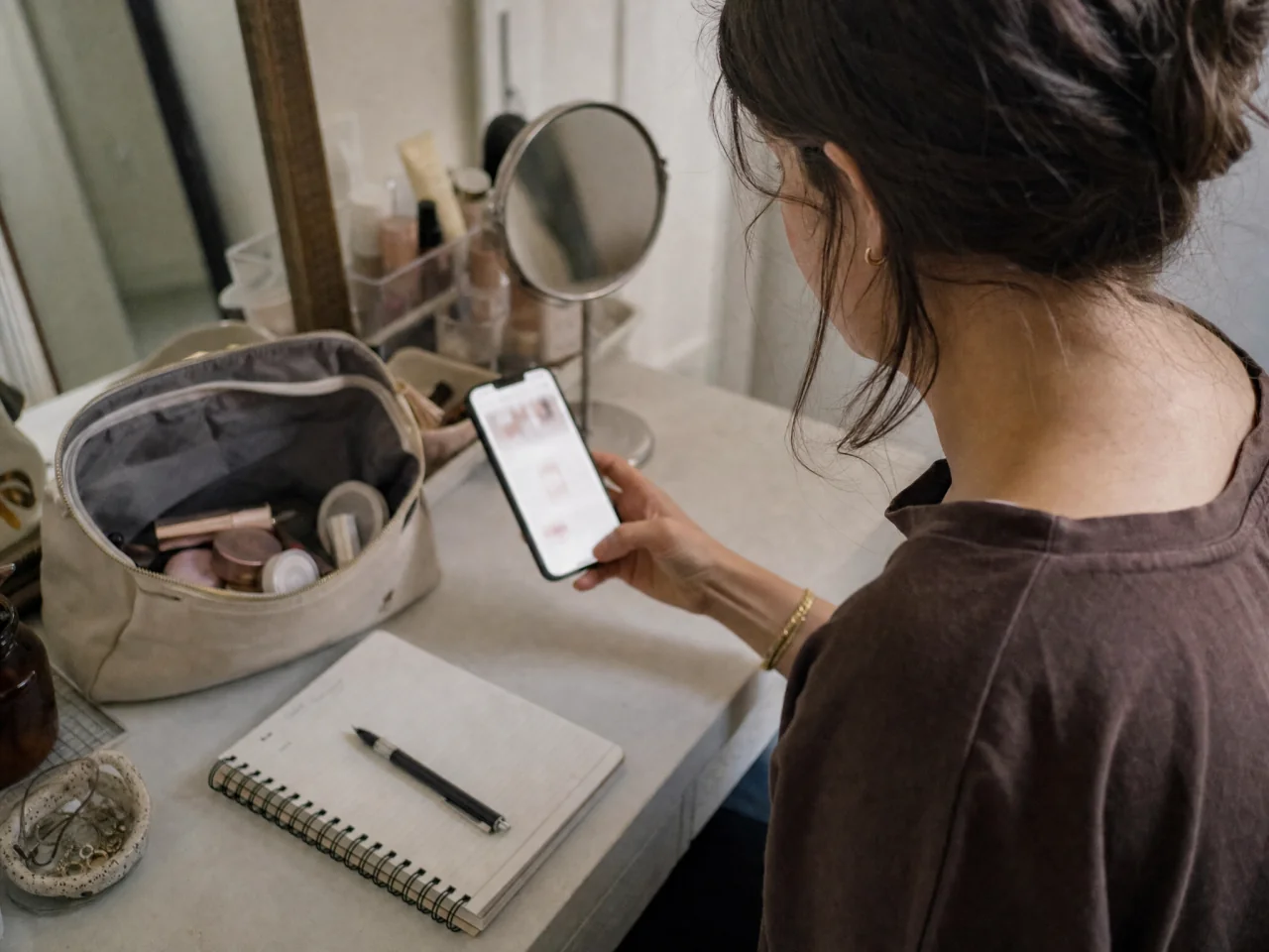 Person at a vanity holding a smartphone with a small open makeup pouch and a notepad nearby, planning a simple natural makeup routine.