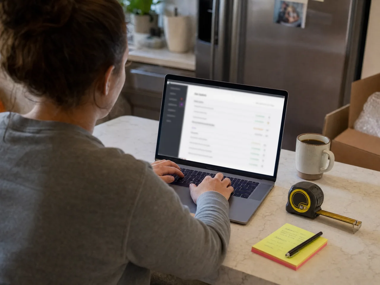 Over-the-shoulder view of a person typing on a laptop with a tape measure and sticky notes on the kitchen counter.